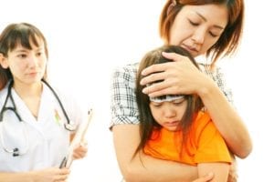 Child in pain from fluoride supplements with patch on her head in mother’s arm with doctor wearing stethoscope looking on